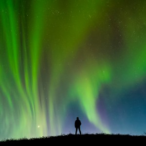 A silhouetted man stands on a hilltop seeming enveloped by the green and purple northern lights or aurora borealis of Southern Iceland.